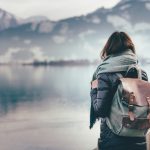 Hipster young girl with backpack enjoying landscape standing near the lake.